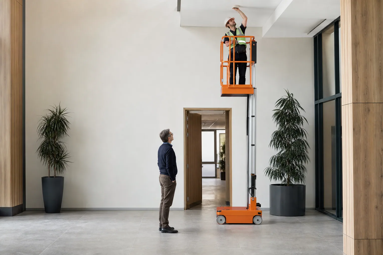 Un technicien intervient au plafond depuis une nacelle compacte à mât vertical dans un atrium.