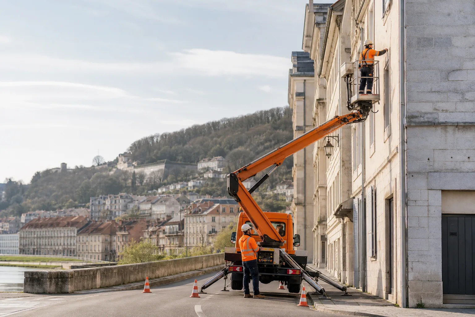 Une nacelle élévatrice orange intervient sur un bâtiment avec un technicien dans le panier et un opérateur au sol, dans le secteur de Besançon.
