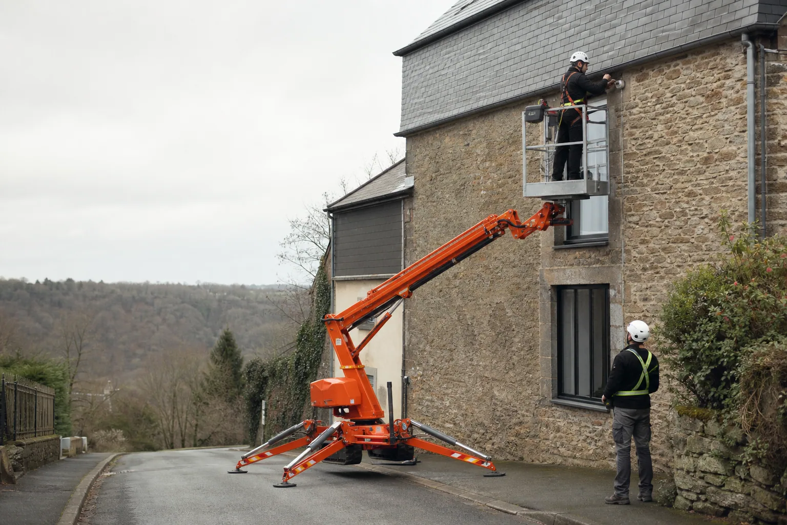 Une nacelle élévatrice orange intervient sur un bâtiment avec un technicien dans le panier et un opérateur au sol, dans le secteur de Fougères.