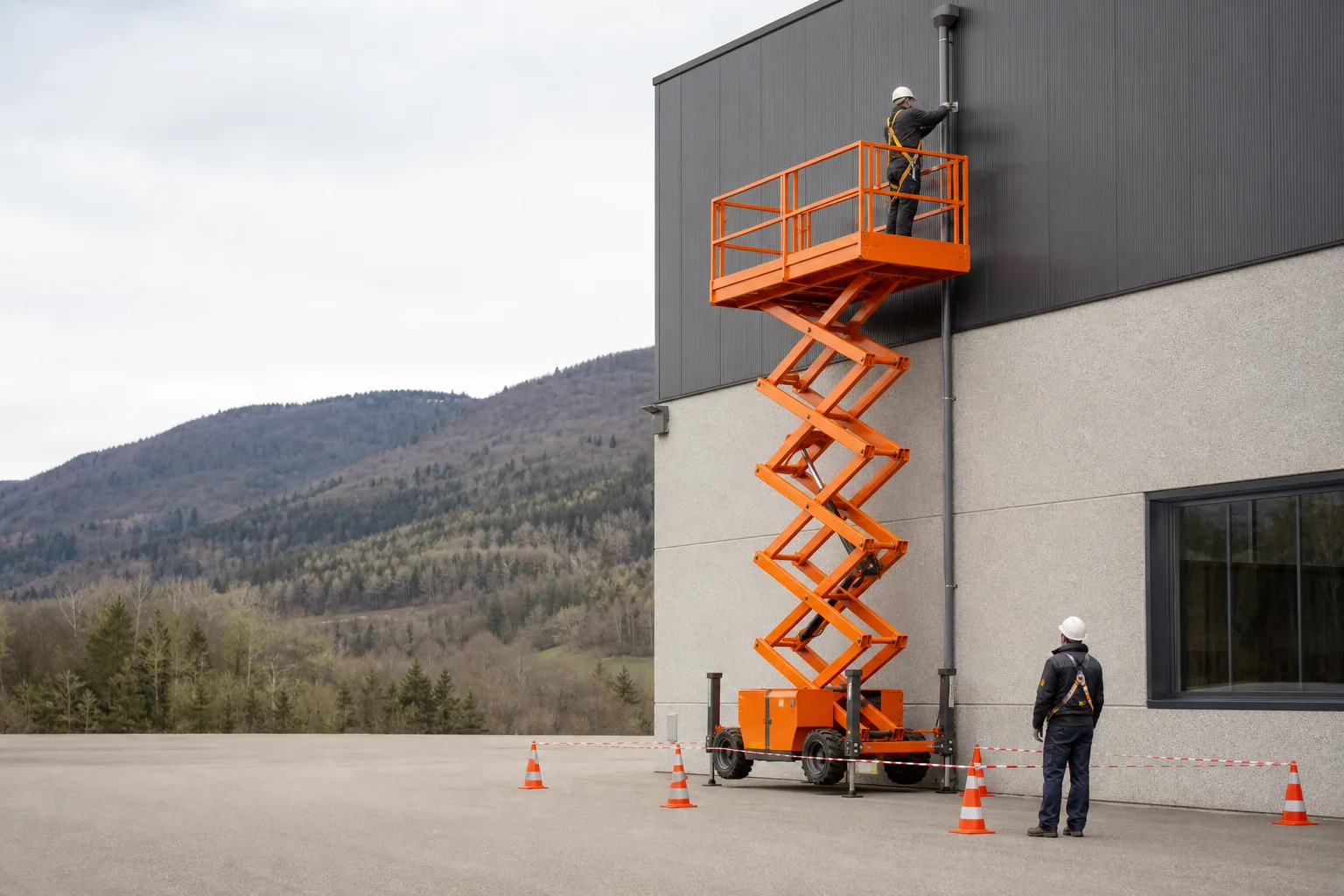 Une nacelle élévatrice orange intervient sur un bâtiment avec un technicien dans le panier et un opérateur au sol, dans le secteur de Oyonnax.